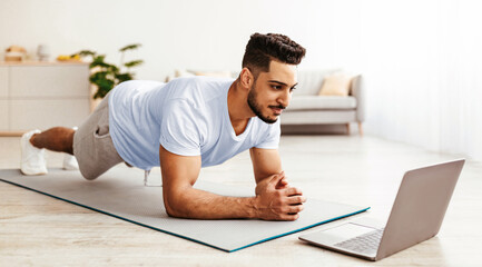 A man is engaged in a plank exercise on a yoga mat at home while following a workout video on a laptop. The room is bright and modern, creating an inviting atmosphere for his fitness routine.