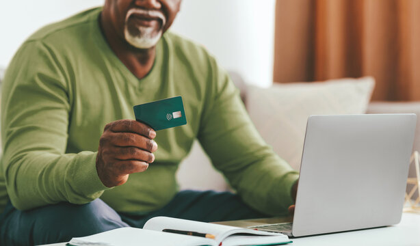 A senior man sits comfortably on a couch, holding a contactless credit card in one hand while using a laptop. He appears focused on his online task, enjoying a relaxed moment at home.