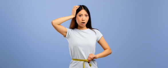 A woman with long black hair looks shocked as she checks her waist measurement with a tape measure. She wears a light blue shirt and stands against a plain blue background.