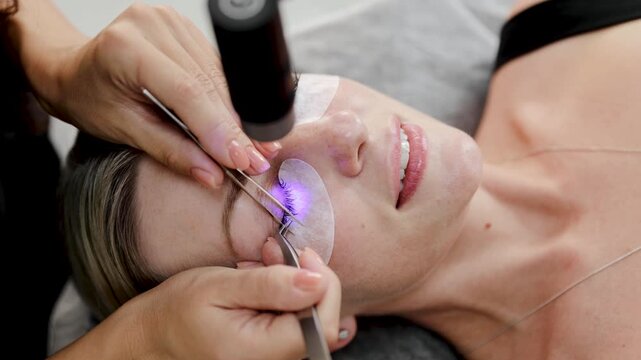Cosmetologist applying false eyelashes with tweezers and curing device in beauty salon