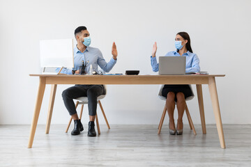 Diverse colleagues wearing disposable face masks are seated at a desk in an office. They wave hands to greet each other, maintaining social distance after self-isolation protocols.