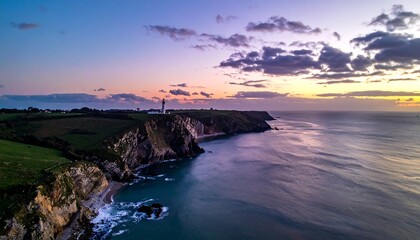Coastal cliffs meet calm ocean under a twilight sky, with a distant lighthouse silhouette