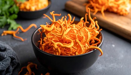 Bowl of vibrant orange cordyceps mushrooms on a gray surface, with other servings on a wooden board