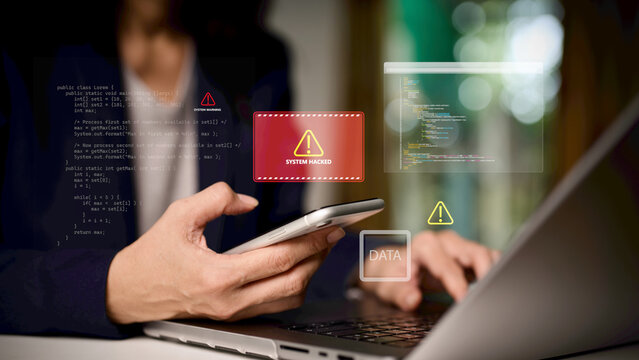 Cybersecurity and data breach concept. Businessperson holding smartphone and working on laptop with digital warning alert “System Hacked”, symbolizing hacking, cyber attack, online data protection.