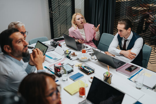 A diverse team collaborates around a conference table filled with laptops, documents, and snacks. The speaker in a pink blazer leads a lively brainstorming session with animated gestures.