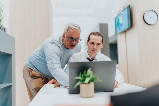 An experienced mentor guides a junior colleague at a bright, clean desk as they work on a laptop. Focused teamwork and professional collaboration drive productivity in a contemporary office.