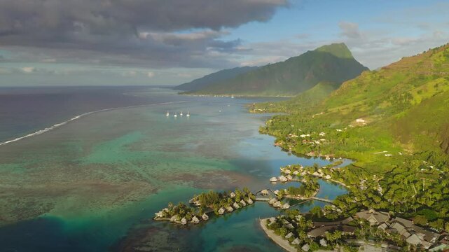 Moorea island showing luxury overwater bungalows on a turquoise lagoon, a fringing reef protecting the coastline, and lush green tropical mountains reaching toward a cloudy sky
