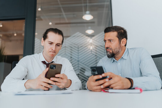 Two diverse professionals sit at a clean desk, focused on their smart phones. The scene conveys modern office work, collaboration, and digital communication in a contemporary business environment.