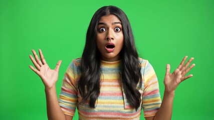 A young Indian woman with long dark hair wearing a striped tshirt stands against a vibrant green screen expressing shock and surprise with wide eyes and open mouth her hands raised in a gesture of di.