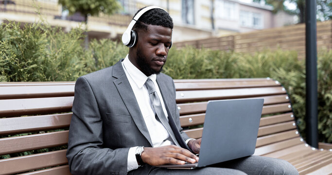 A businessman in a suit sits on a bench in a city park. He focuses on his laptop while wearing headphones. Surrounding him are green bushes and a modern urban setting.
