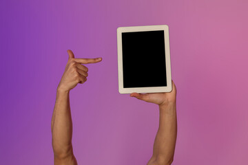 Cropped view shows a millennial man pointing at a blank tablet screen illuminated by colorful neon lights. This setup is ideal for showcasing mobile app designs or online advertisements.