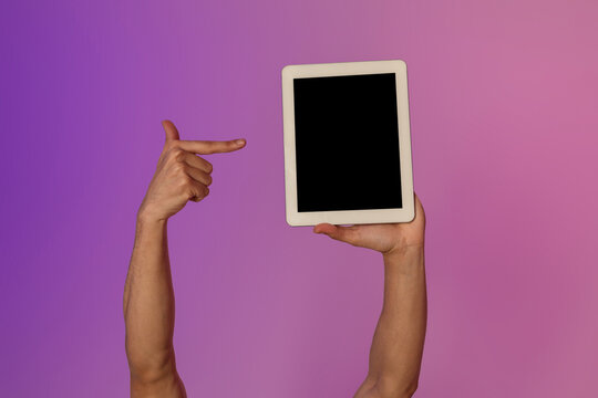 Cropped view shows a millennial man pointing at a blank tablet screen illuminated by colorful neon lights. This setup is ideal for showcasing mobile app designs or online advertisements.