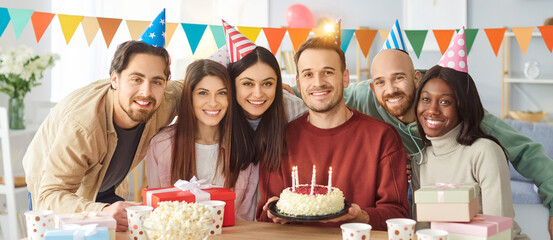 Photo of cheerful diverse friends celebrating special occasion, enjoying birthday party, posing in decorated room at table with cake and gift boxes, enjoying celebration in merry company