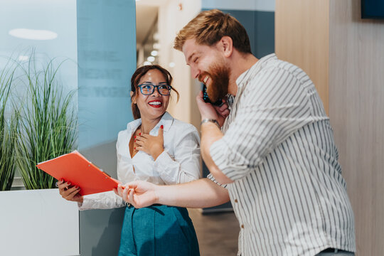 Two coworkers in a modern office laugh and chat as one holds an orange folder and the other speaks on the phone, conveying collaboration, positivity, and teamwork.