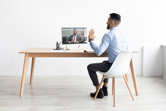 A young Arab man sits at a desk in an office, smiling and waving at a colleague on a video call. He is engaged in remote work, using a laptop for the conference.