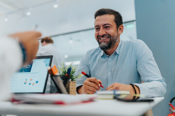 Smiling bearded man works at a bright, modern office desk, writing with a red marker beside a laptop and charts. The scene conveys teamwork, planning, and a productive multicultural business setting.