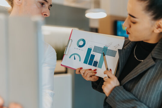 Two professional women review a data-filled clipboard, analyzing graphs and metrics in a collaborative office setting. The scene emphasizes teamwork, analytics, and diverse leadership.