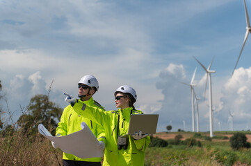 Engineers wearing safety helmets inspecting a wind turbine at a wind farm and holding a blueprint while working.