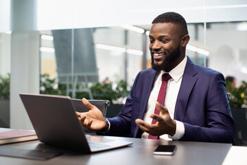 Happy black businessman in expensive suit sitting at table in office, having video chat with...