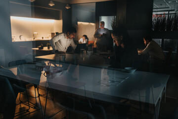 A group of professionals gathers around a glass conference table in a modern office, dimly lit with warm accents. Reflections and a focused, collaborative atmosphere suggest after-hours work and