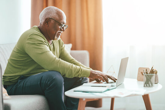 An elderly man sits comfortably on a sofa using his laptop. The bright morning light filters in, creating a warm and inviting atmosphere. He appears focused and engaged in his work. - Powered by Adobe