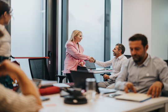 A professional office scene shows a smiling woman in a pink blazer shaking hands with a man at a desk, while colleagues work nearby, conveying collaboration and corporate teamwork.