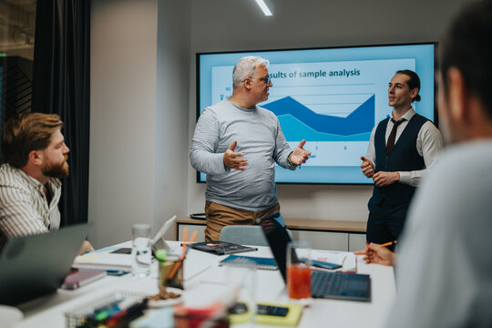 A team briefing in a modern office with a presenter and a speaker beside a large display showing a chart, while teammates work on laptops and take notes.
