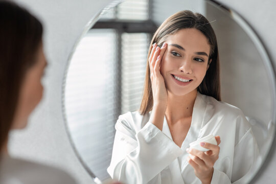 Attractive young female smiles at her reflection while applying moisturising cream in a bright room. She enjoys her skincare routine and appreciates her skin's condition.
