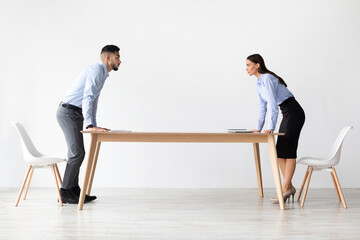 Two young professionals, a man and a woman, confront each other in a modern office. They lean over a table, expressing disagreement and anger in a tense work situation.