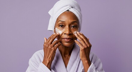 Mature woman with a towel on her head applies eye cream in a bathrobe against a purple background