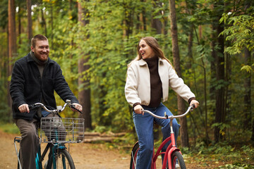Obraz premium Caucasian young man and Caucasian young woman riding bicycles through forest path, smiling and looking at each other, surrounded by green trees, enjoying outdoor activity together