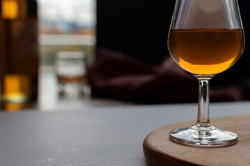 Elegant glass of amber brandy or cognac on a wooden stand in a dim, atmospheric bar or tasting room. A bottle and blurred background create a sophisticated mood