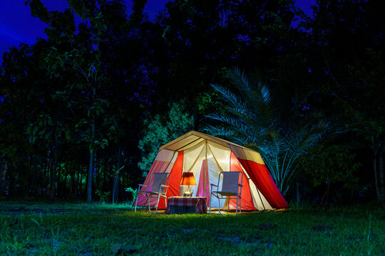 Camping tents at night, vintage cabin tents, antique oil lamps, retro chairs, and a group of camping tents with outdoor coffee-making facilities on wooden tables in a forest camping area.