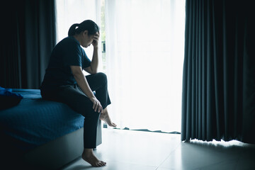 A stressed woman sitting alone on bed in dark room, holding her head down, symbolizing depression, anxiety, hopelessness, loneliness, mental health struggle, burnout and emotional pain.