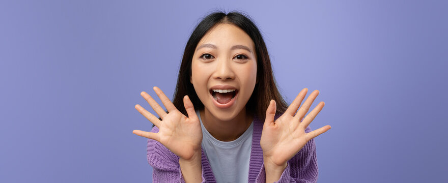 A young woman with long hair smiles widely while raising her hands in excitement. She wears a purple sweater and a shirt, standing in front of a solid purple backdrop.