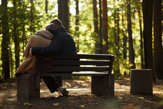 Young Caucasian couple sitting on bench embracing in forest, woman resting head on man's shoulder, both facing away from camera surrounded by tall trees and sunlight