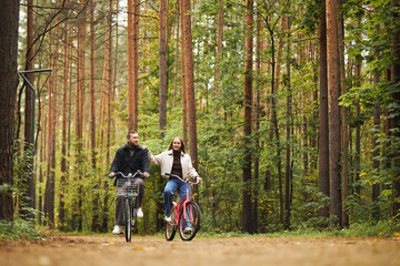 Caucasian young adult man and Caucasian young adult woman riding bicycles together on forest path, smiling and talking, surrounded by tall trees and lush greenery in outdoor setting