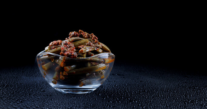 Smoke curls around a clear glass bowl of bracken fern salad with spices. Fern salad on a black background.