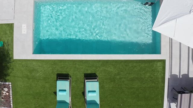 Top view of a turquoise rectangular swimming pool with two sunbeds and a white parasol on a tiled patio surrounded by artificial turf in a private villa