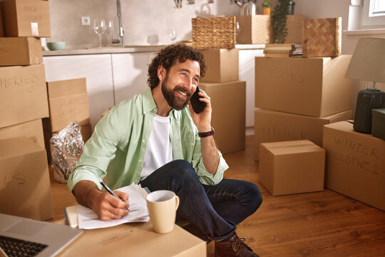A man sits on the floor amidst unpacked boxes, holding a phone to his ear while jotting down notes in a notebook. His relaxed demeanor suggests he is coordinating moving details.
