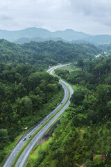 Aerial view of a winding road curving through lush green tropical forest with misty atmosphere. Scenic mountain highway surrounded by dense jungle and natural landscape.	
