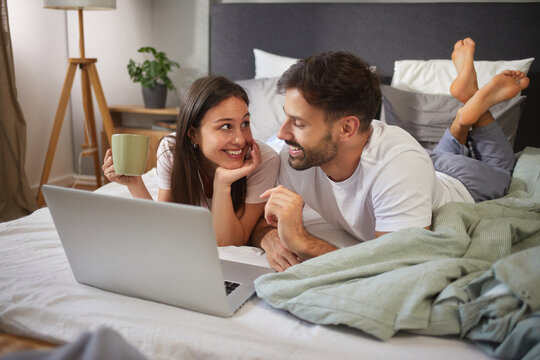 A young couple relaxes together in bed, sharing smiles and ideas while using a laptop. The warm atmosphere is enhanced by soft bedding and a cup of coffee, creating an intimate morning moment.
