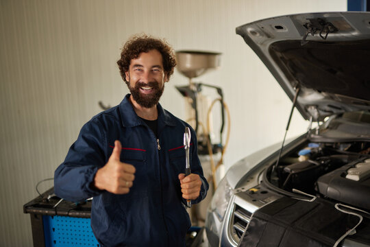 A skilled car mechanic is smiling and showing a thumbs up while holding a wrench. He is working under the hood of a car in a bright repair shop.