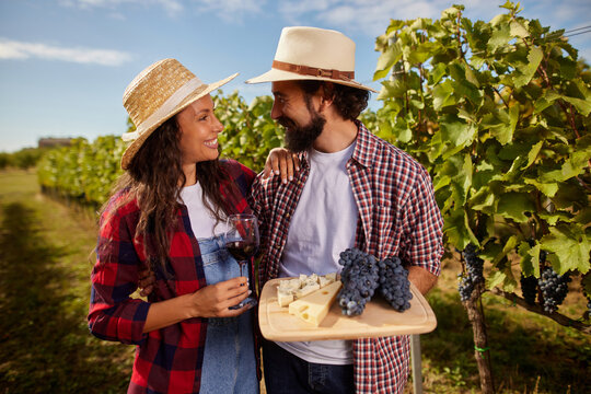 A couple smiles at each other while enjoying a moment in their family vineyard. They hold a glass of red wine and a platter of cheese and grapes amidst lush vines on a sunny day.