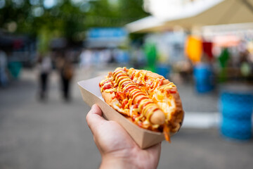 A close-up, point-of-view shot of a hand holding a delicious hot dog topped with ketchup and mustard in a paper tray at an outdoor food market © pavel siamionov