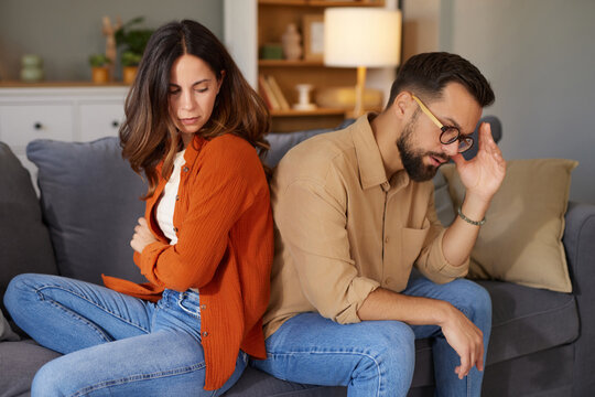 A woman and a man sit on a sofa, facing away from each other, expressing tension and unresolved issues. The living room is warmly decorated, highlighting their emotional distance.
