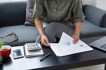 Businesswoman sitting at desk on couch in workplace or at home working on laptop and analyzing data on charts and graphs and writing on papers to make business plan and strategies for company