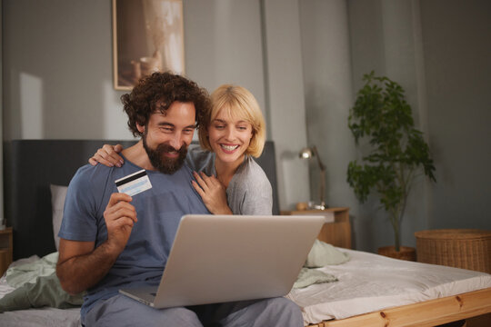 A man and woman are happily shopping online from their bed, using a credit card and a laptop. The room is warmly lit, creating a relaxed atmosphere.
