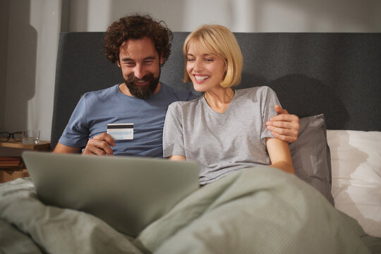 A couple relaxes in bed, engaging in online shopping on a laptop while sharing smiles. The cozy room provides a warm and inviting atmosphere as they spend quality time together. - Powered by Adobe