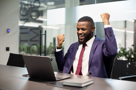 Emotional wealthy black businessman sitting in front of laptop at modern office, looking at computer screen and raising hands up, celebrating success in his business, copy space